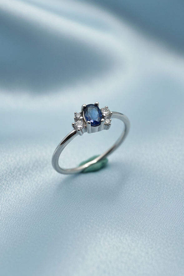 A silver ring with a central oval blue gemstone and small clear accent stones, photographed on a plain light background.