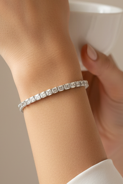 A sparkling tennis bracelet set with clear stones in a silver-toned band, held in a person's hand for scale. A laptop and patterned fabric are visible in the background.