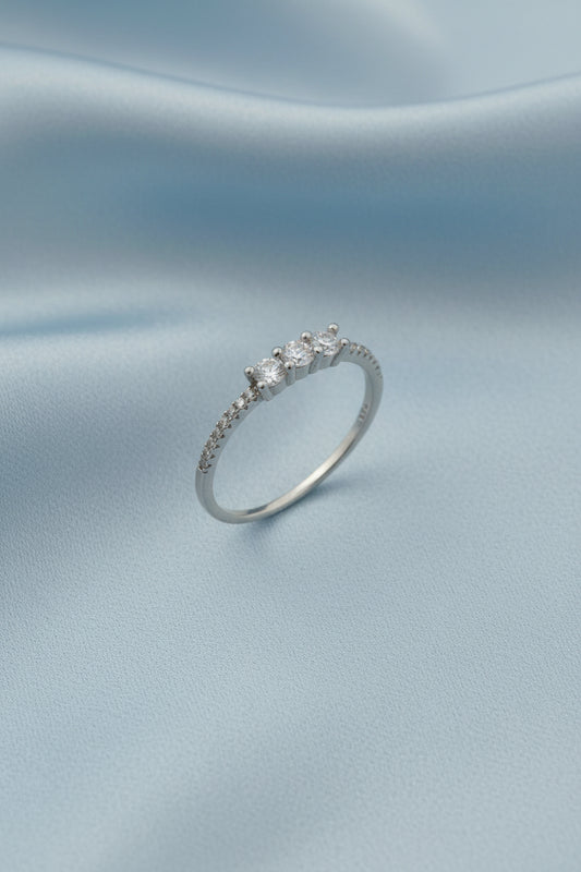 A silver band ring featuring three round-cut clear gemstones in a row, with small pavé-set accents along the shoulders, displayed on a light gray reflective surface.