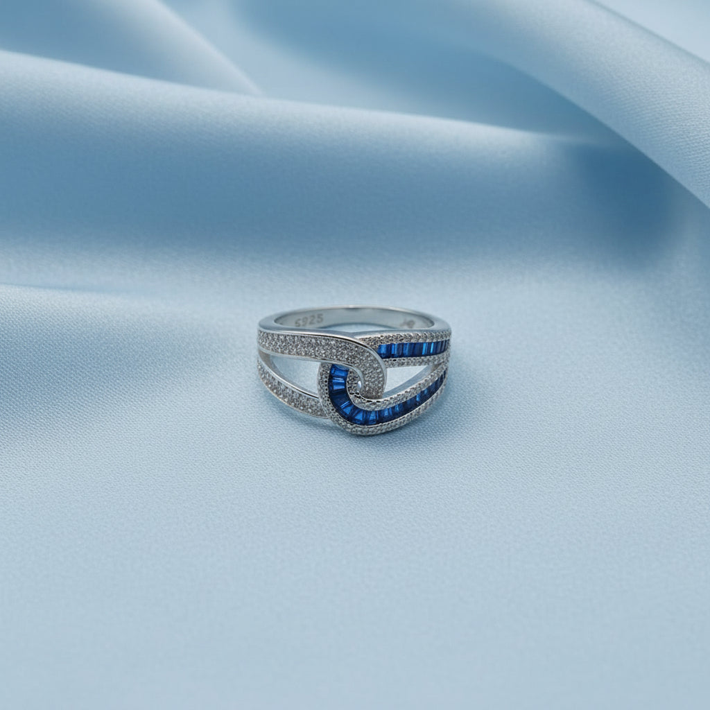 A silver-toned ring with a double-curve design, featuring inlaid and channel-set deep blue gemstones and small clear stones, photographed on a light gray background.
